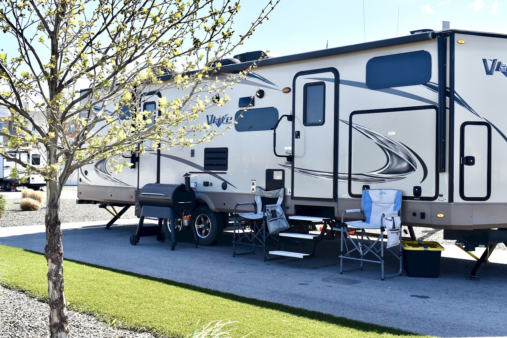A parked RV with outdoor chairs, a grill, and a small table set up on a paved driveway next to a tree.