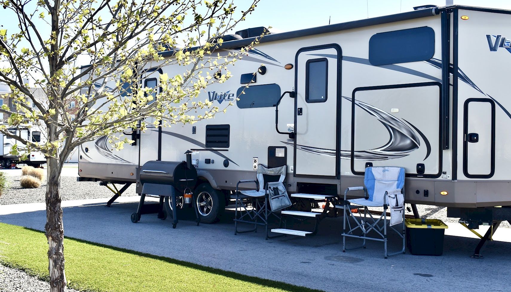 A parked RV with outdoor chairs, a grill, and a small table set up on a paved driveway next to a tree.