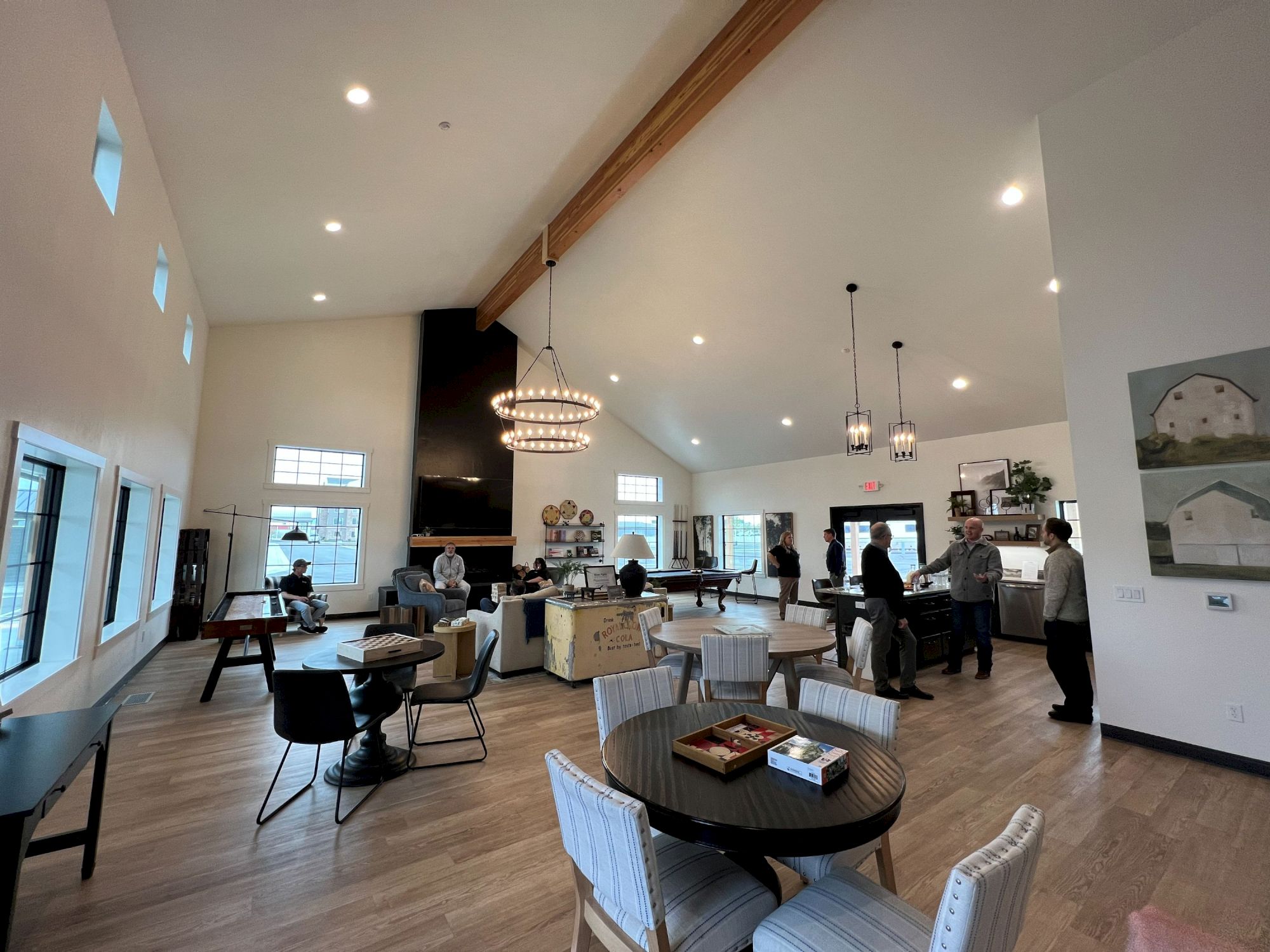 People dining in a bright, modern cafe with round tables, hanging lights, wooden floors, and large windows.