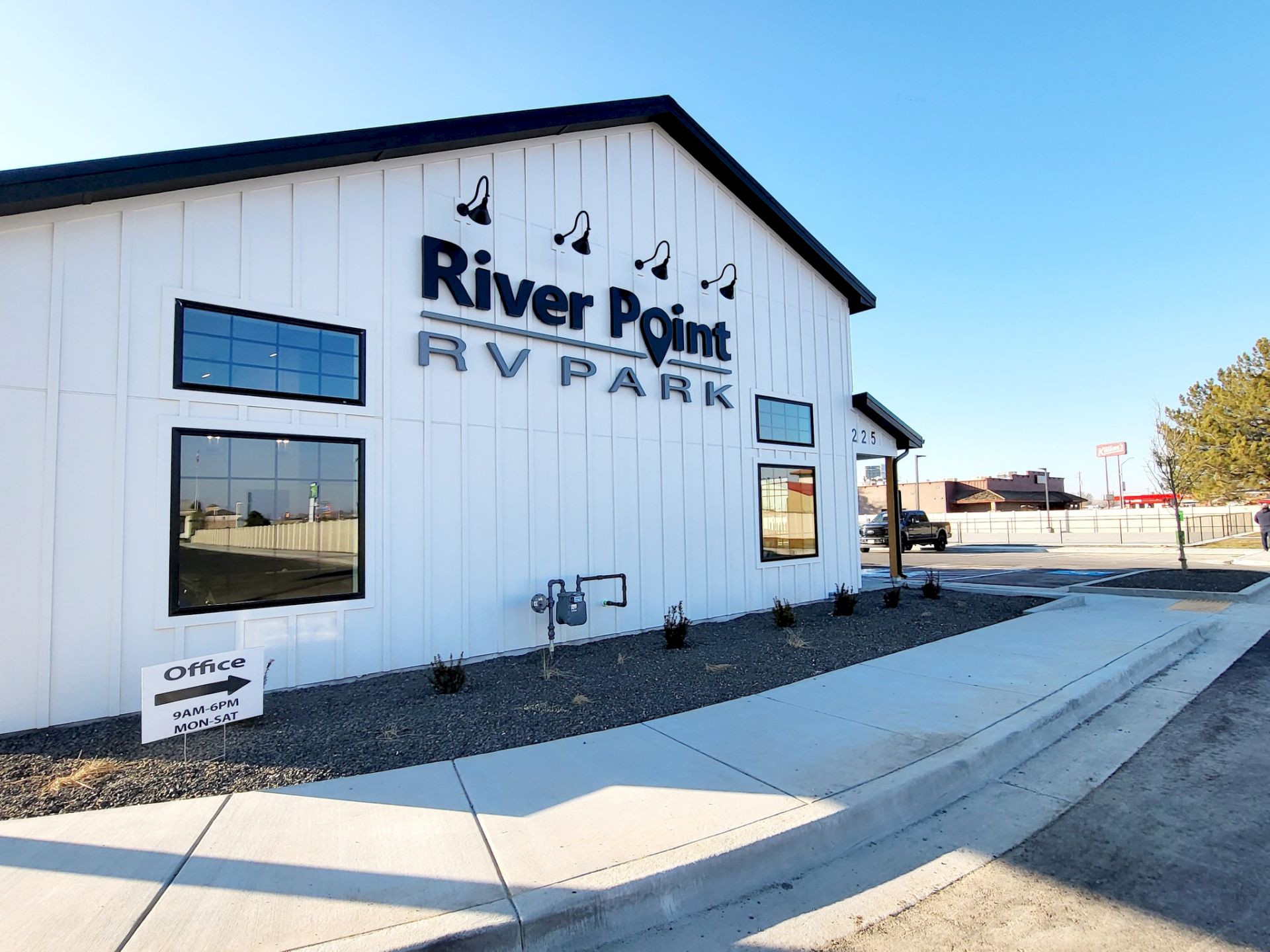 River Point RV Park building with sign, white siding, glass windows, landscaped curb, clear blue sky, and a wide sidewalk in front.