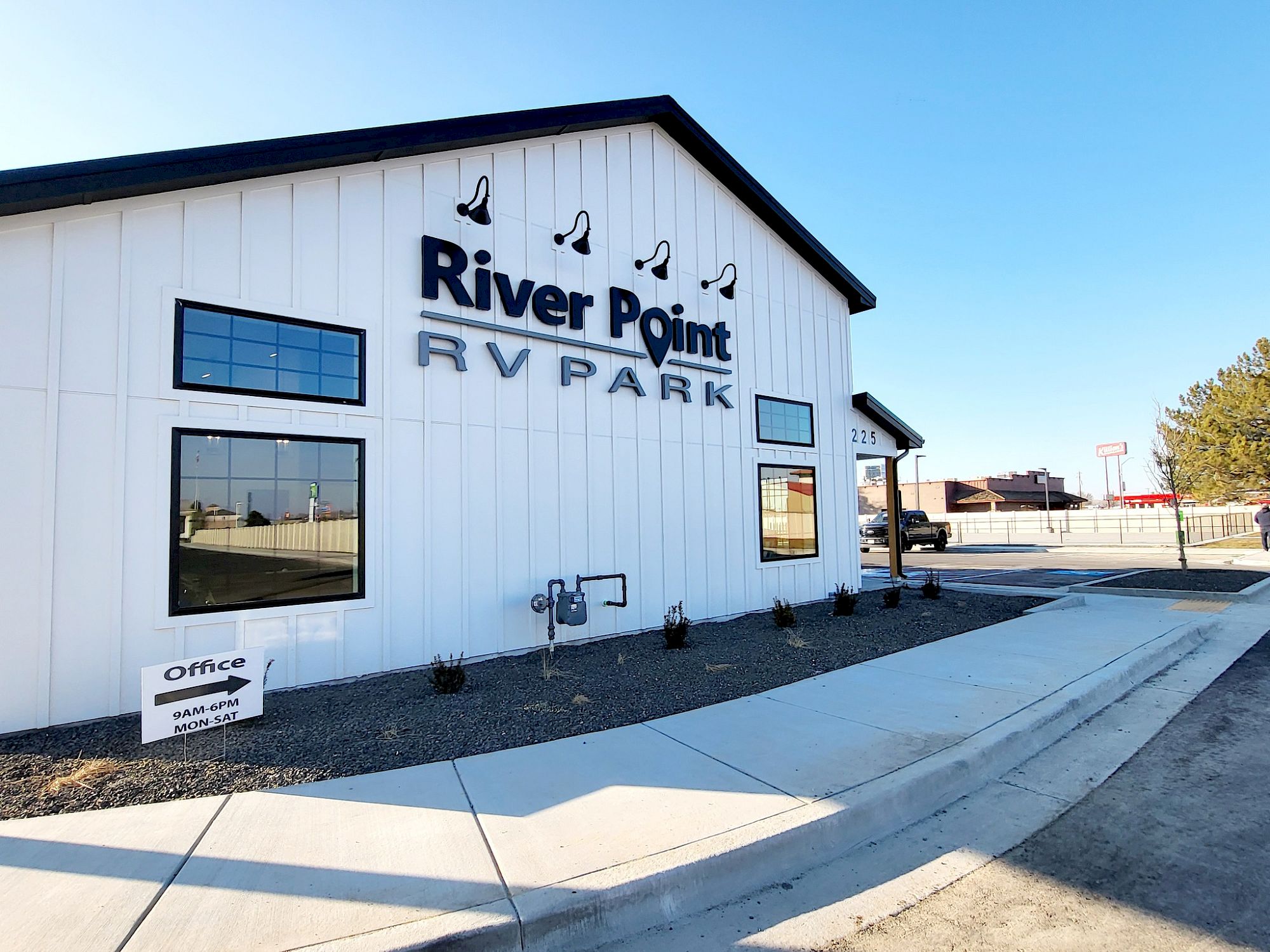 River Point RV Park building with sign, white siding, glass windows, landscaped curb, clear blue sky, and a wide sidewalk in front.