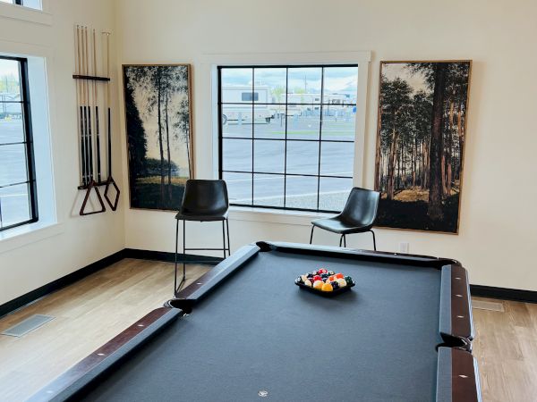 A billiards room with a pool table, two chairs, wall art, and large windows letting in natural light.