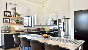 A modern L-shaped kitchen with white cabinets, stainless steel appliances, a large island with a checkered countertop, and bar seating, ending with a door.