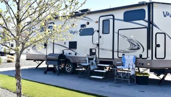 A parked RV with an outdoor setup: chairs, a grill, and a small table on a paved space beside a tree under sunny skies.