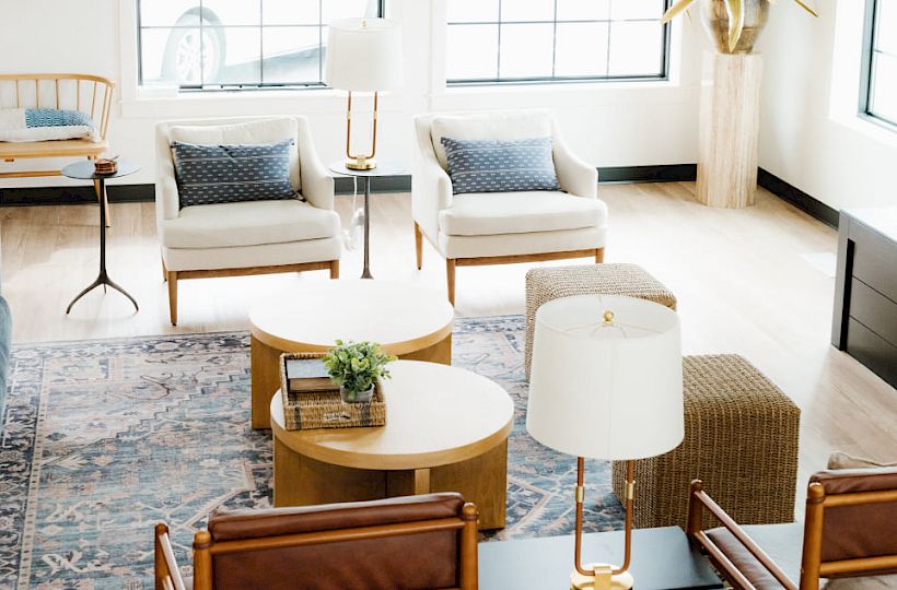A bright, airy living room with mid-century chairs, a dark coffee table, a patterned rug, tall windows, and a modern chandelier.