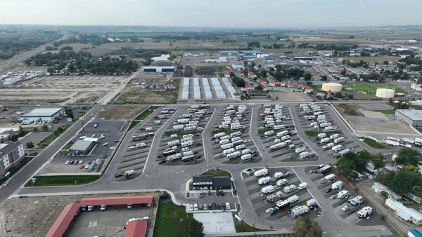Aerial view of a large RV park or marina area with rows of campers, vehicles, and buildings, plus open fields and distant industrial facilities.