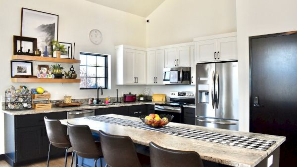 A bright modern kitchen with white cabinets, stainless steel appliances, a long marble island, bar seats, and decorative shelves on the wall.