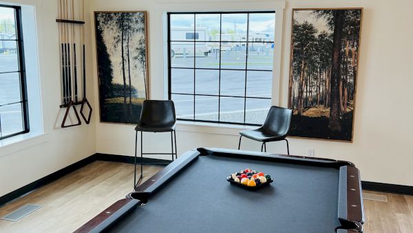 A modern game room with a pool table centered under a window, two black chairs, and nature-themed vertical art on the walls, bright and airy.