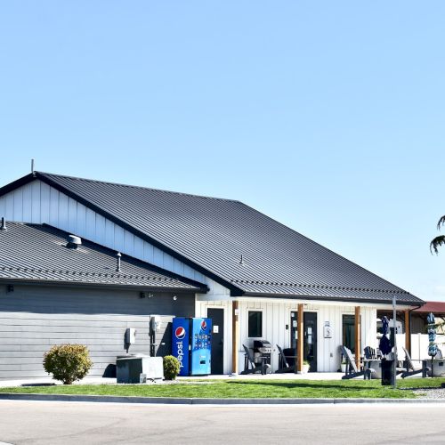 A modern, single-story building with a gray roof, a two-car garage, a small front lawn, and vending machines near the entrance, under a clear blue sky.