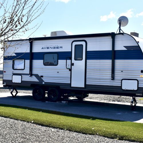 A white travel trailer with blue stripes sits on a paved pad, parked beside a grassy area under a clear sky.