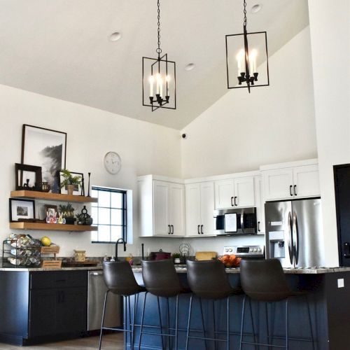 A modern white kitchen with an island, black bar stools, pendant lantern lights, and stainless appliances.