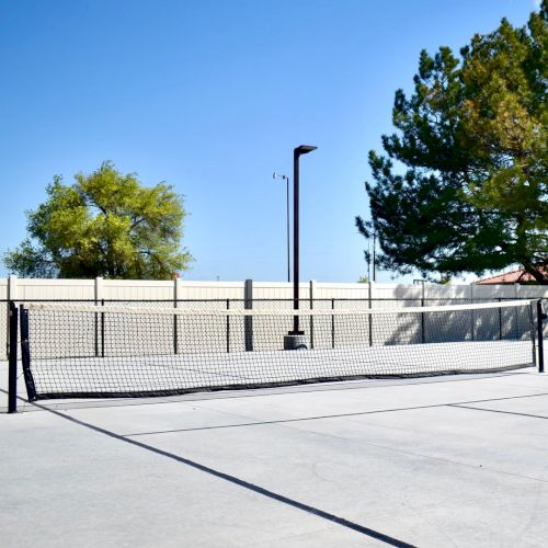 An outdoor fenced tennis court with a net, concrete ground, and trees under a bright blue sky.