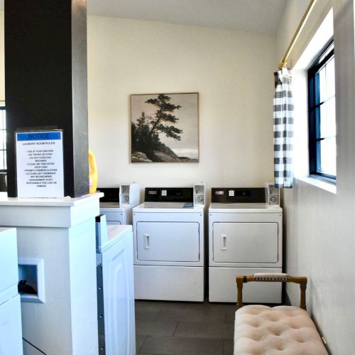 A laundry room with multiple dryers and a washing machine, a window with a checkered curtain, and a small cushioned bench.