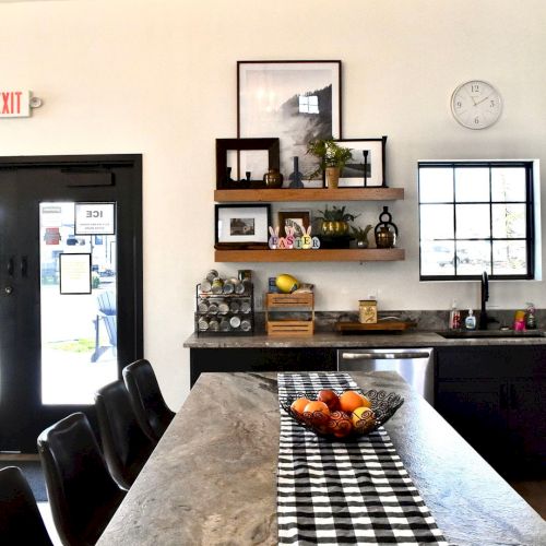An office/kitchen area with a long dining table, black-and-white runner, wooden shelves, Easter decor, clock, and window.