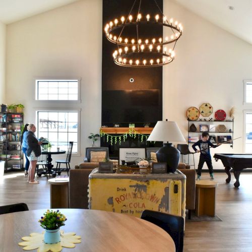 Bright living room with a large candle chandelier, kids by a piano, and a dining table in the foreground.