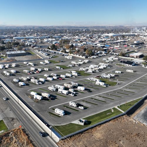 A vast RV park with rows of white trailers, some shade structures, and surrounding roads under a clear sky, with a few trees and open ground.