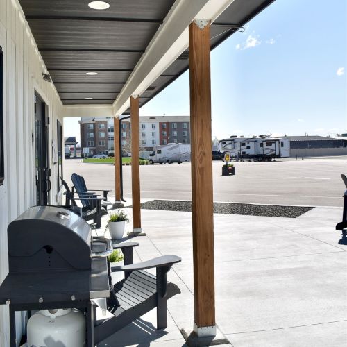 A covered patio with a charcoal grill and chairs, overlooking a parking lot with an RV and other vehicles outside.