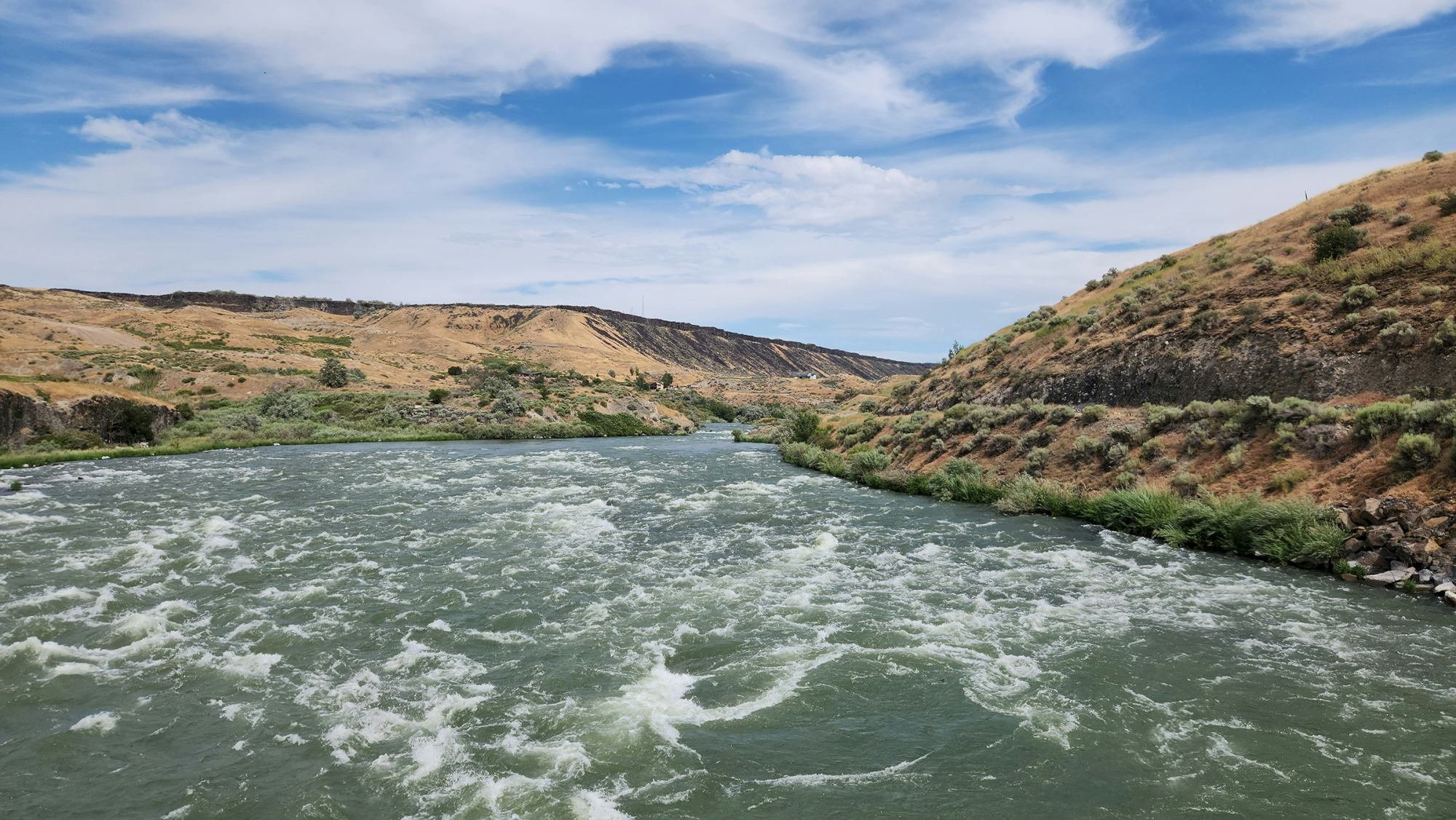 A winding river cuts through arid, grassy hills under a bright blue sky with wispy clouds and rocky slopes along the banks.