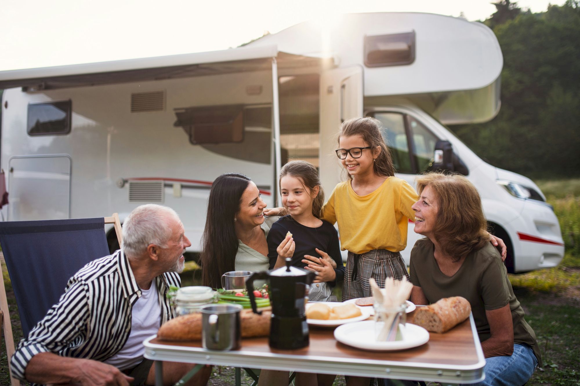 A family sits at a fold-out table outside a camper van, sharing a cheerful meal with coffee cups and plates in a sunny outdoor setting.