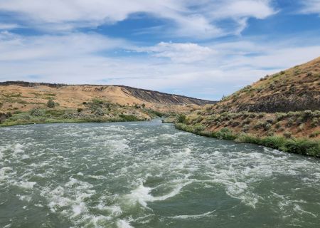 A wide, winding river cuts through rocky hills with sparse vegetation, under a bright blue sky with scattered clouds.
