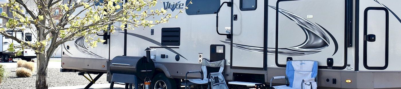 A white travel trailer/RV parked outdoors with chairs and outdoor gear under spring trees.