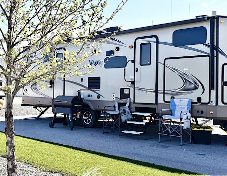 A white travel trailer/RV parked outdoors with chairs and outdoor gear under spring trees.